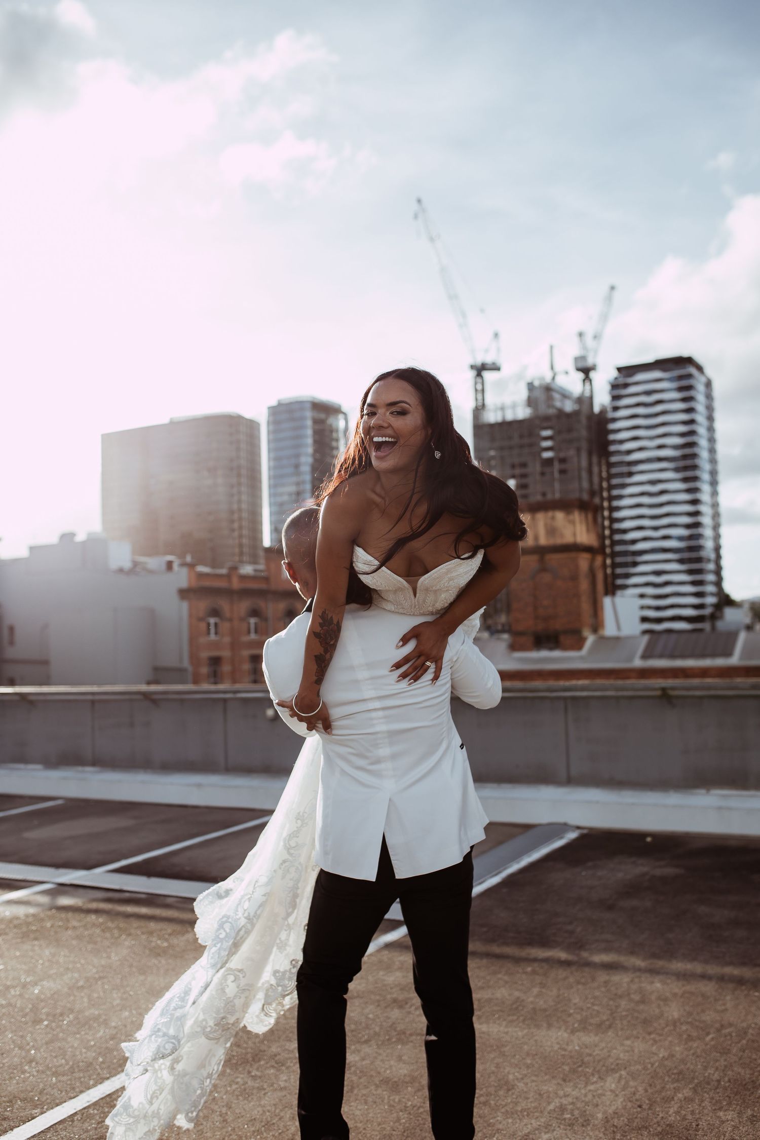 A joyful couple embraces on an urban rooftop with city buildings and construction cranes visible in the background.