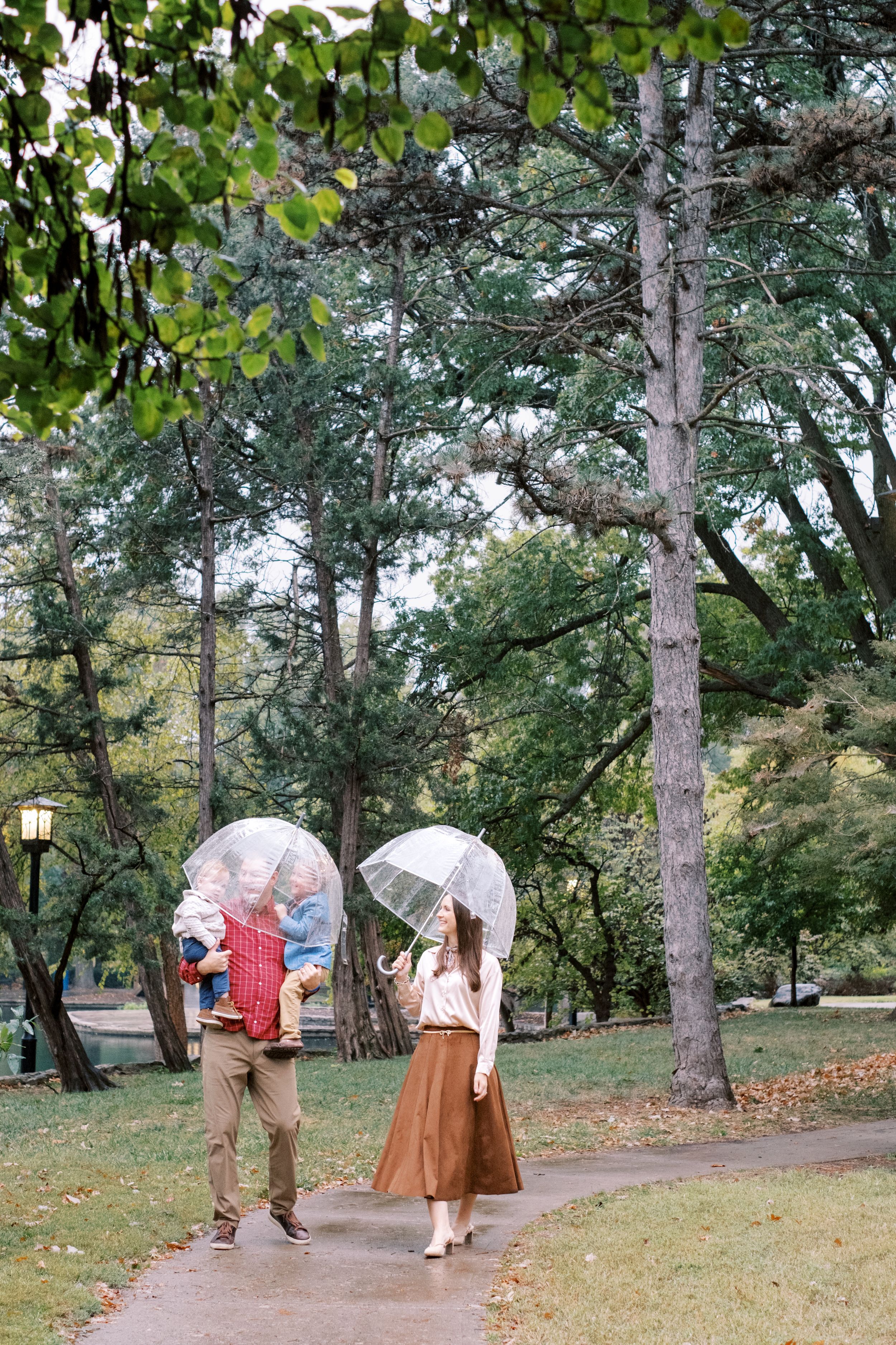 Family walking in a rainy park with clear umbrellas, man holding a child, woman in brown skirt among lush green trees.