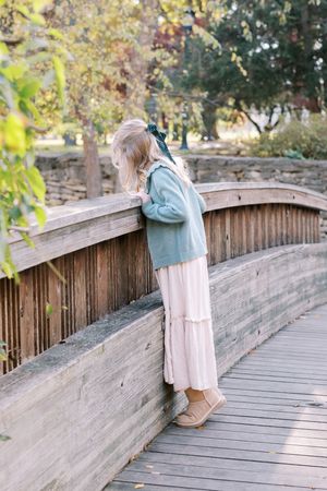 A person in a pink dress and blue backpack stands on a curved wooden bridge walkway in a park setting.