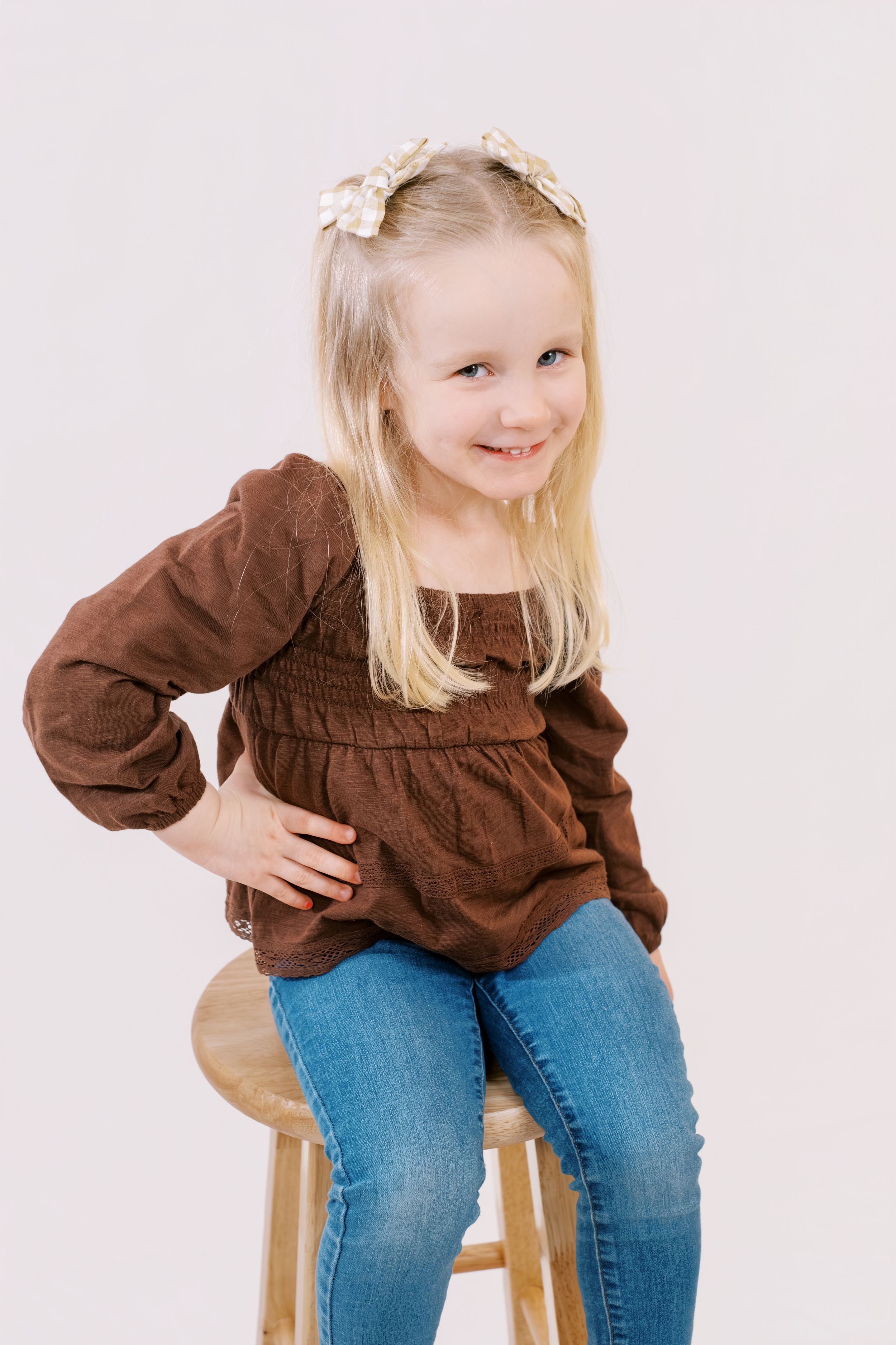 A child in a brown smocked top and jeans sits on a wooden stool against a light background.