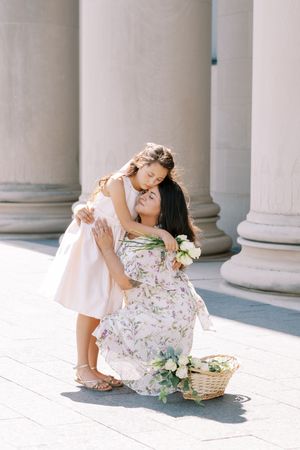 Woman in floral dress bends down with woven basket among classical columns in soft sunlight.