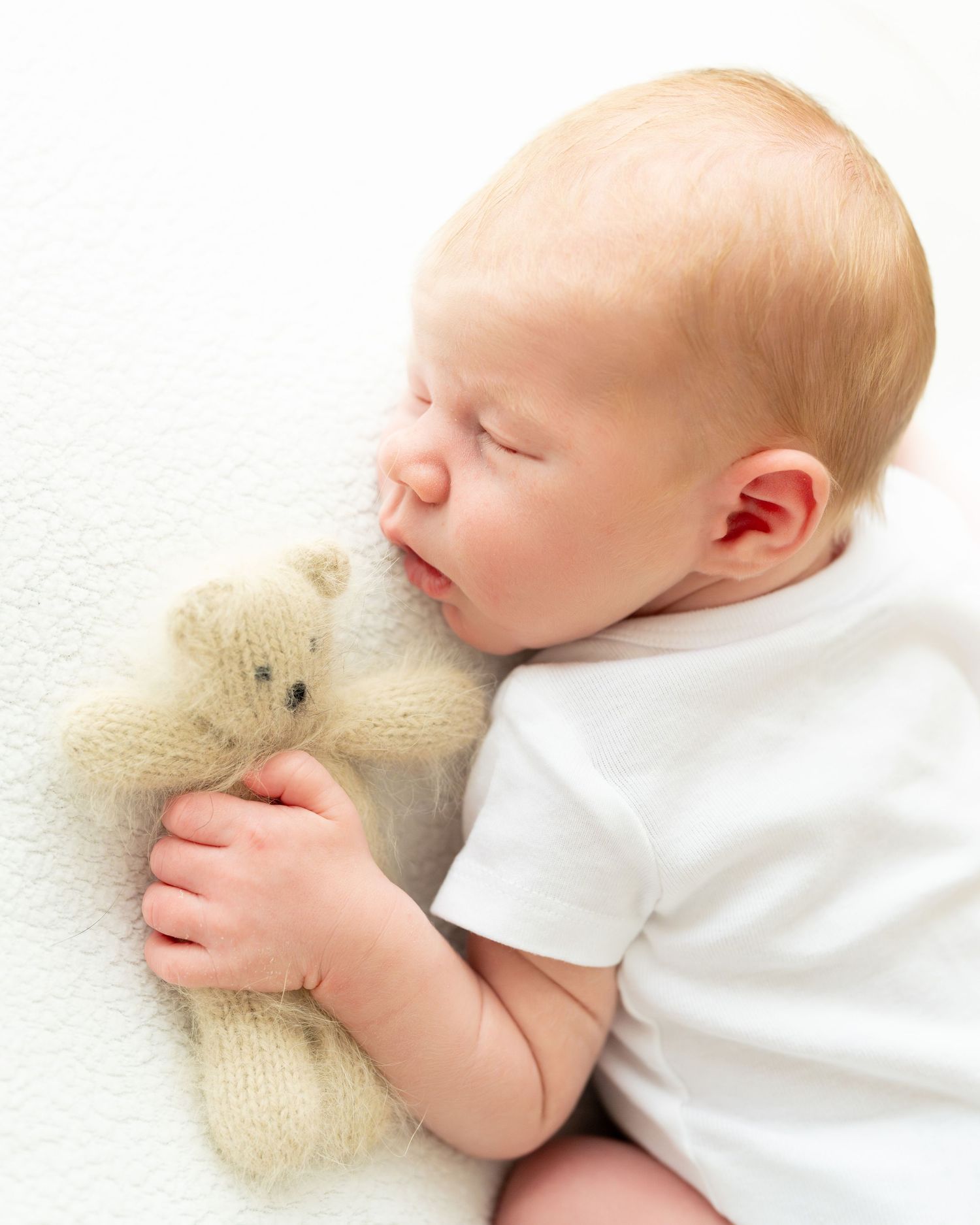 Sleeping baby cuddling with a small teddy bear on white knit blanket in peaceful slumber.