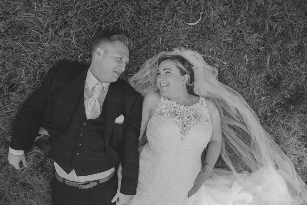 A bride an groom take a lie down on their wedding day, holding hands in a field, captured in a black and white photo