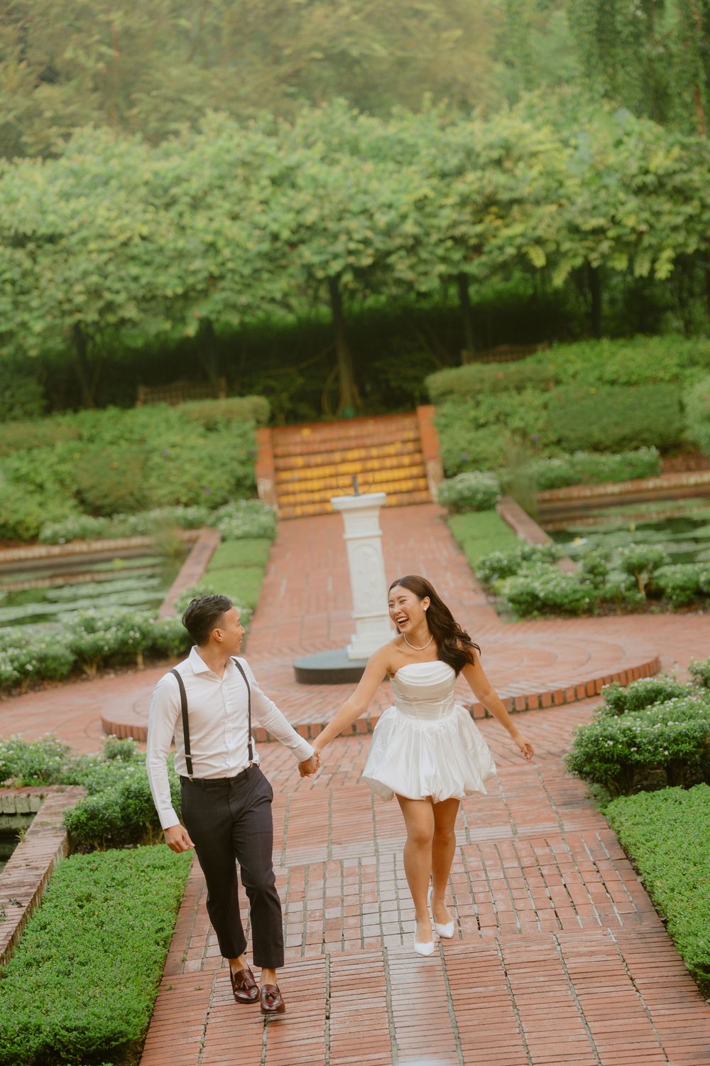 A couple walks hand in hand along a winding brick pathway surrounded by lush green gardens.