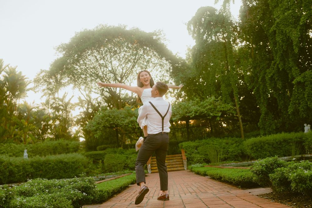 A joyful figure leaps with outstretched arms on a garden path surrounded by lush greenery at sunset.