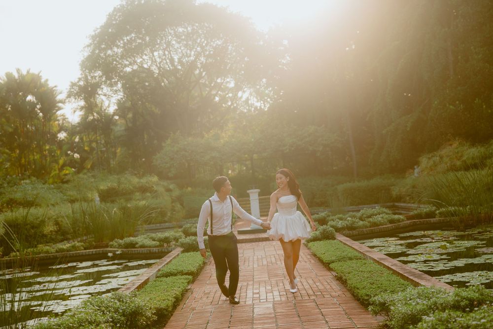 A couple walks hand in hand along a brick path beside a pond during a golden sunset.