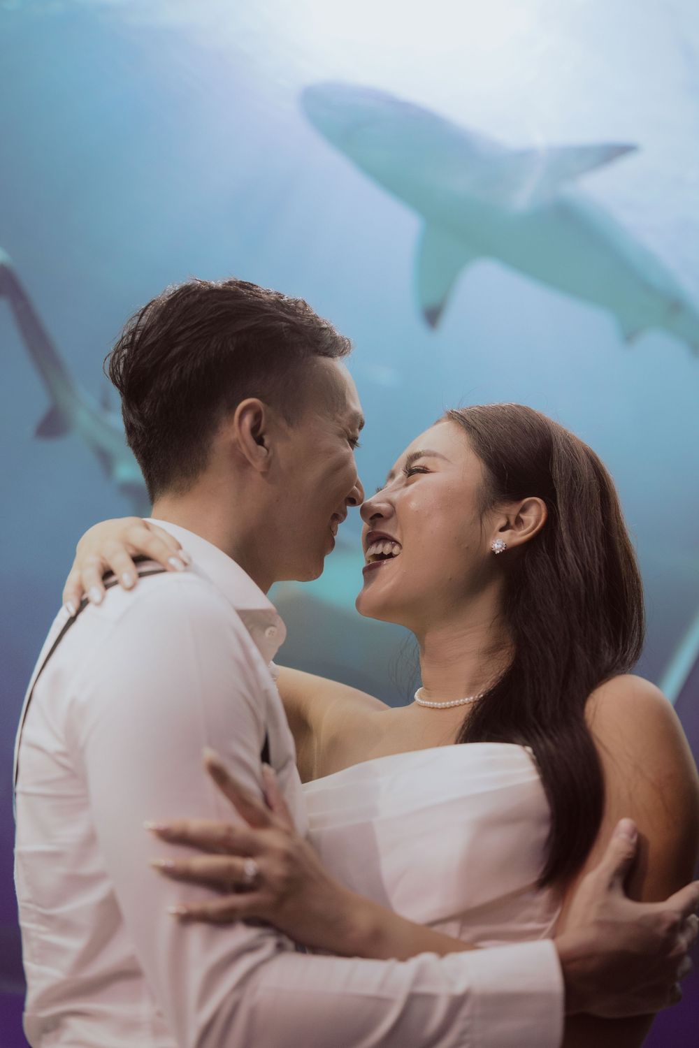 A romantic couple embraces and laughs together while sharks swim in an aquarium tank behind them.
