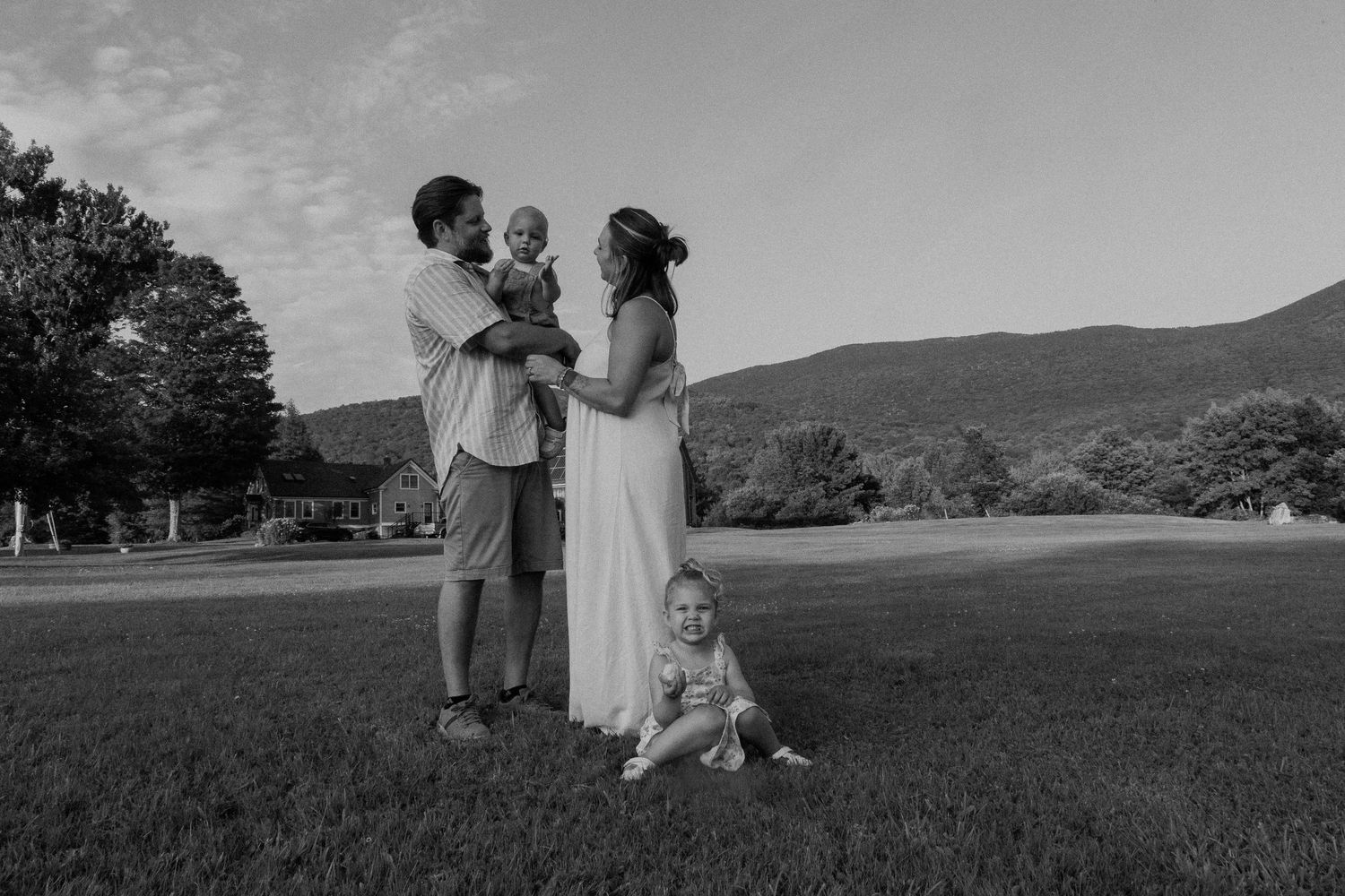Black and white photo of a family relaxing together on grass in a park setting with trees in the background.