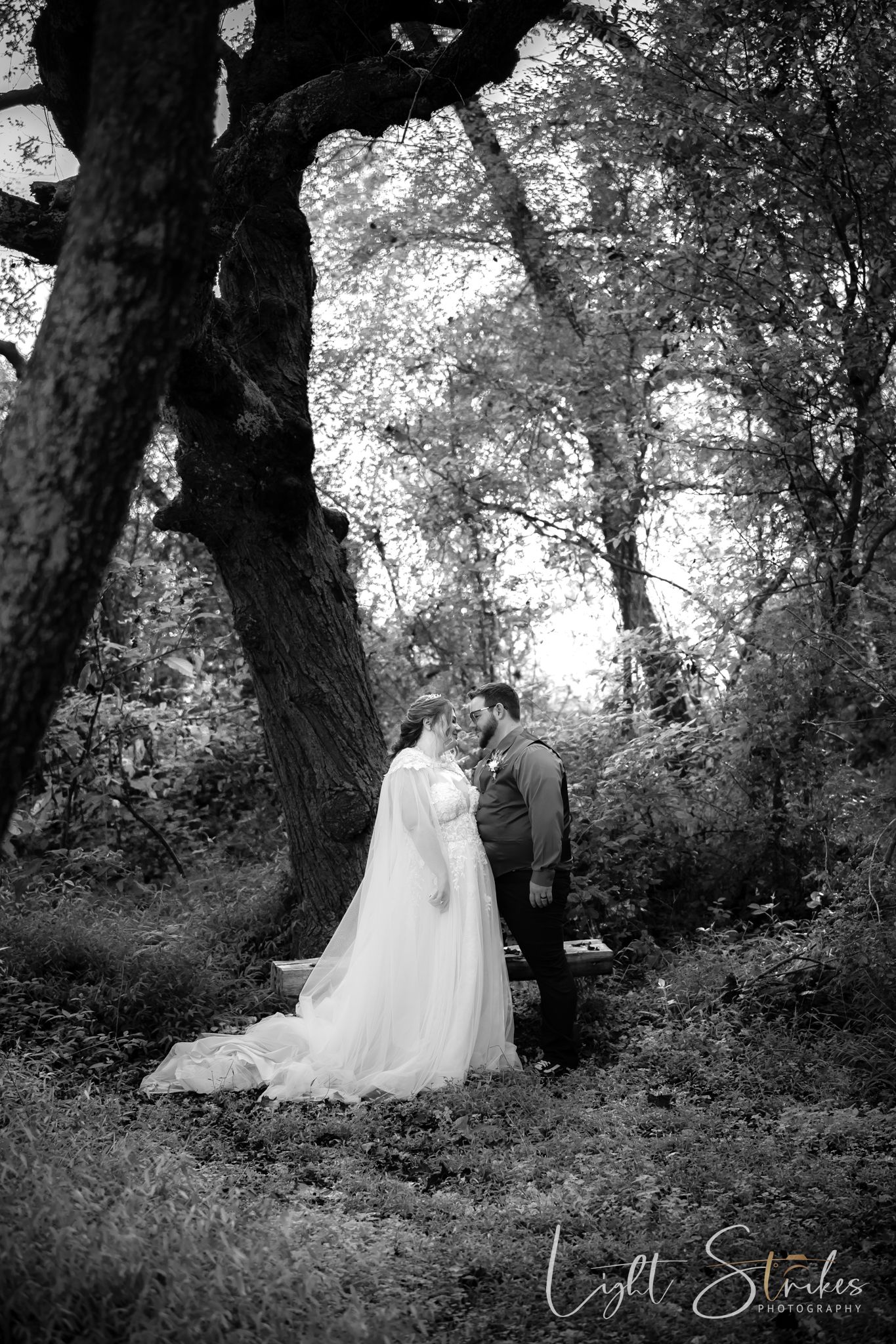 A romantic black and white wedding photo of a couple standing together under tall trees in a forest setting.