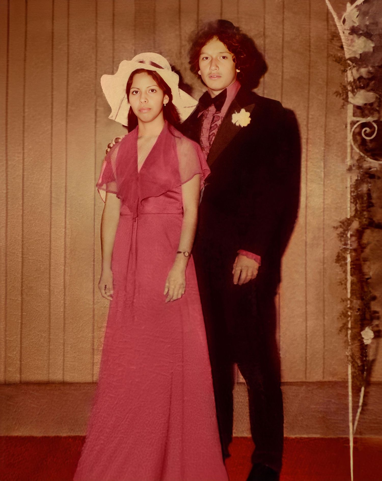 A vintage 1970s formal photograph showing a couple in evening attire - a pink dress and dark suit - against a wood-paneled wall.