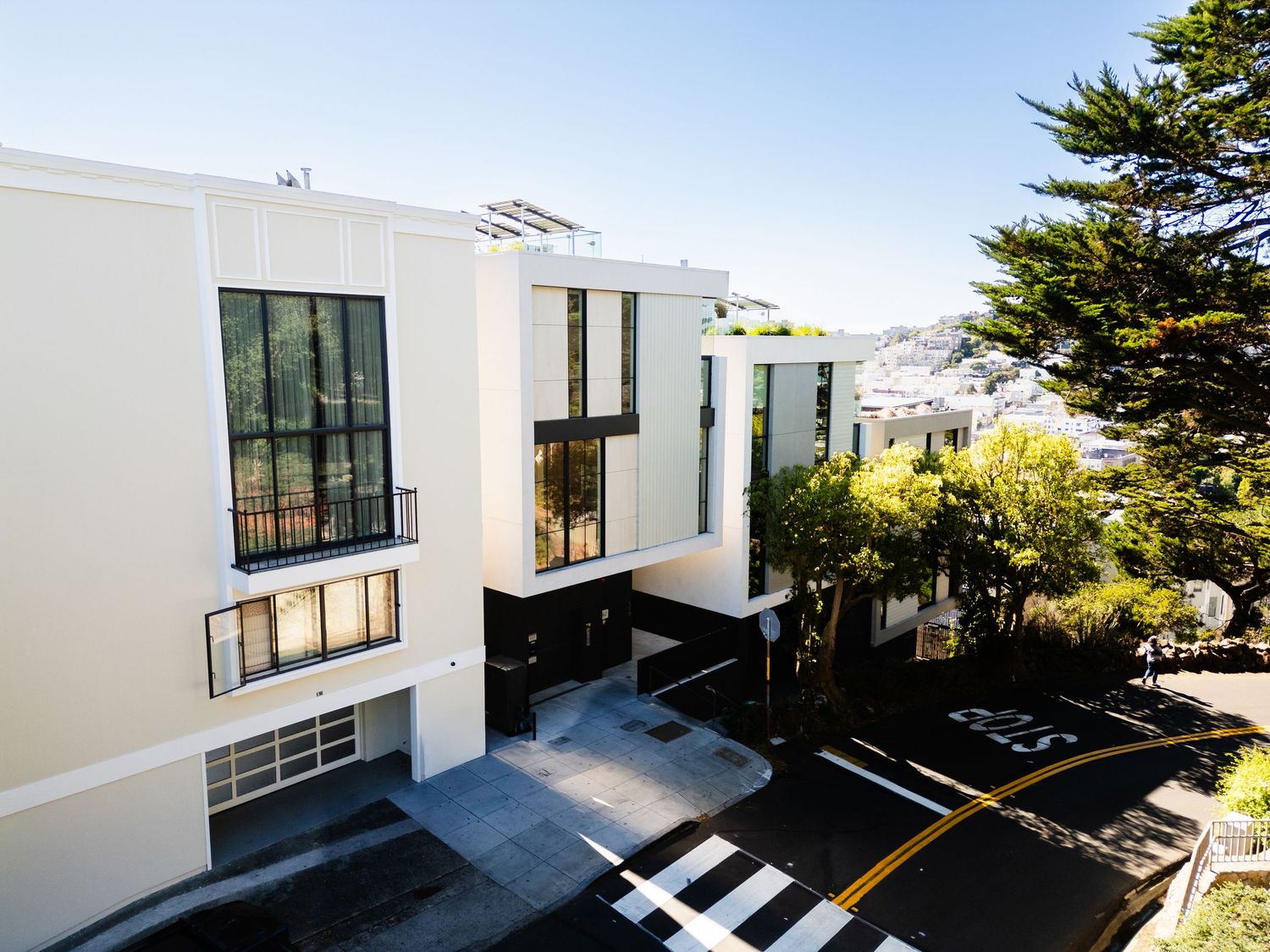 Modern white residential building with large windows and balconies nestled among trees on a hillside.
