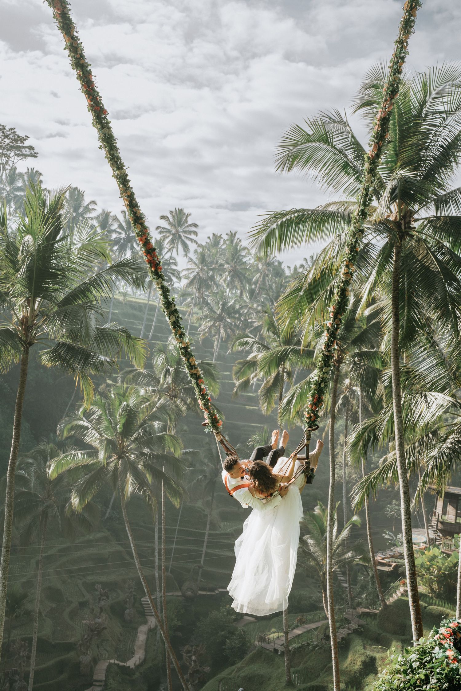 Person in white dress swinging on giant rope swing among tropical palm trees in Bali.