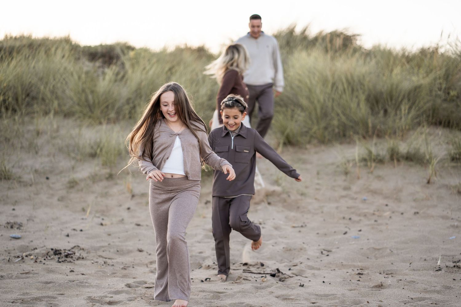 Couple walking together on a sandy beach path through tall grass during sunset.