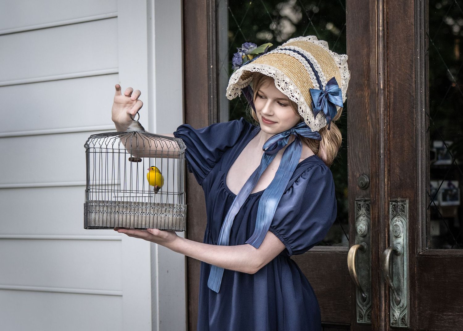 Young woman in blue vintage dress and straw bonnet holds a birdcage with yellow canary near wooden door. Best photographer Williamsburg VA