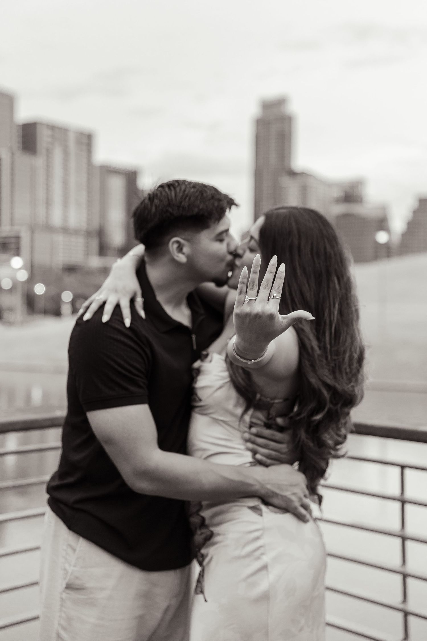 A romantic engagement photo showing a couple kissing while the woman displays her ring against a city skyline backdrop.