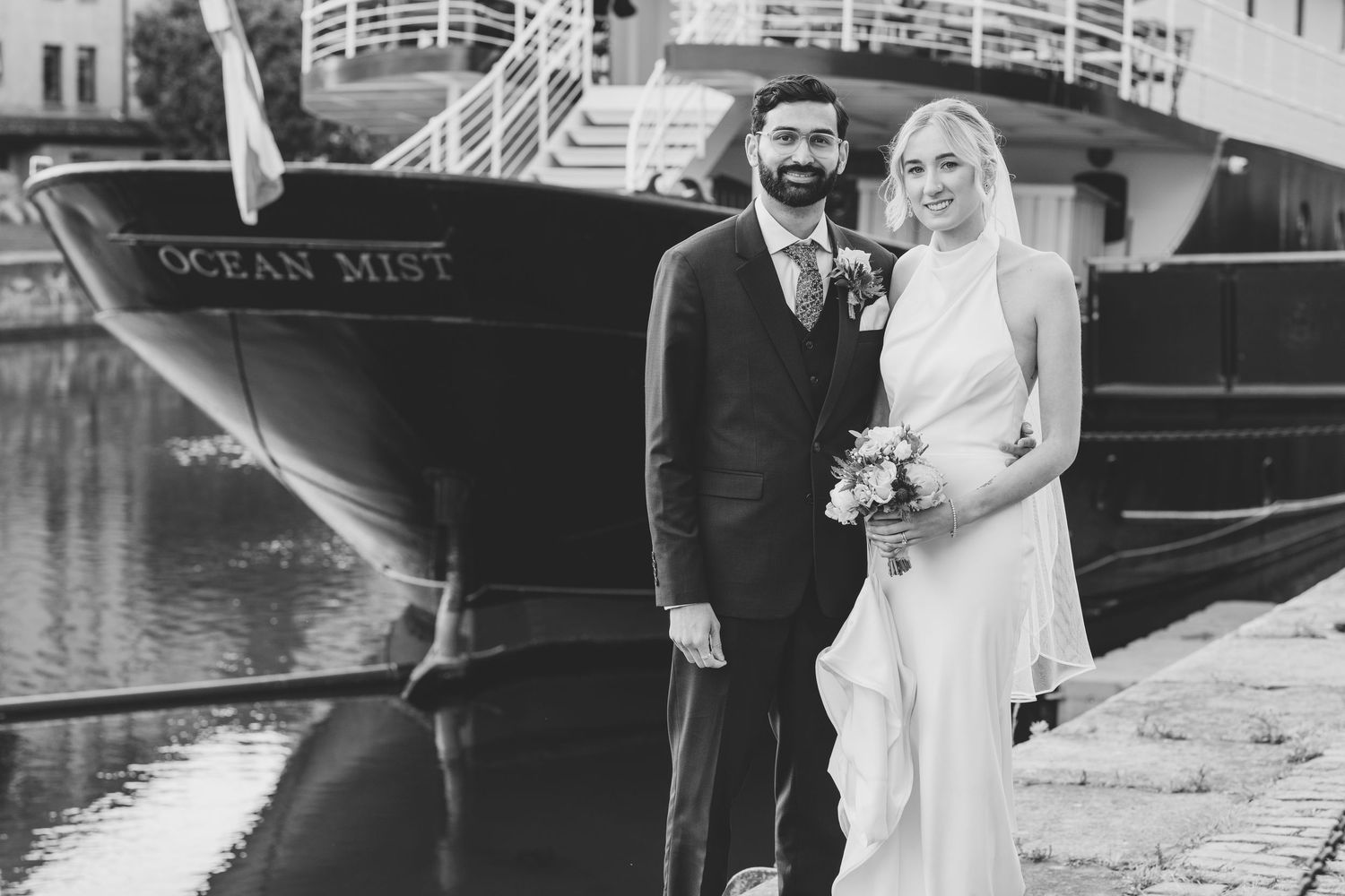 Newlyweds posing by waterfront with docked boats in background.