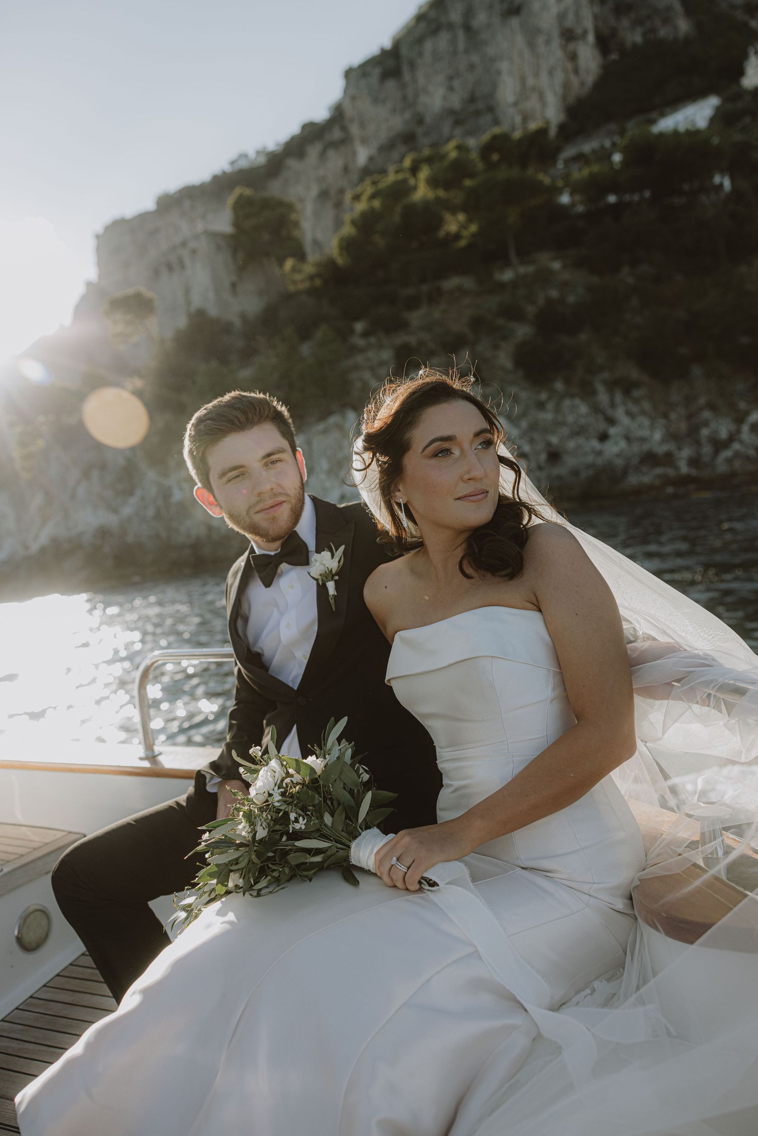 Couple in wedding attire sitting on dock with dramatic coastal cliffs in background.