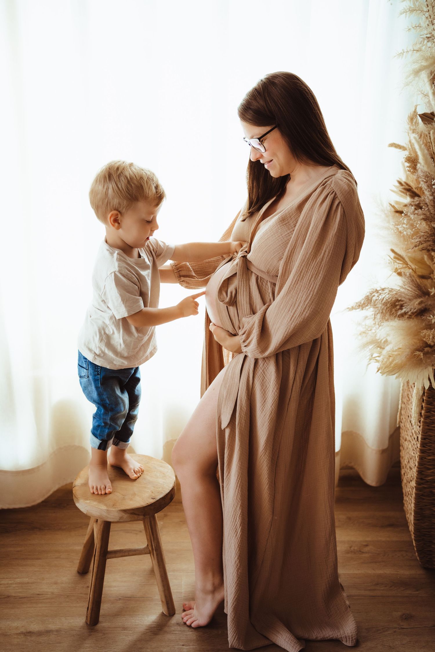 A touching moment between a pregnant woman in a beige dress and a small child standing on a wooden stool.