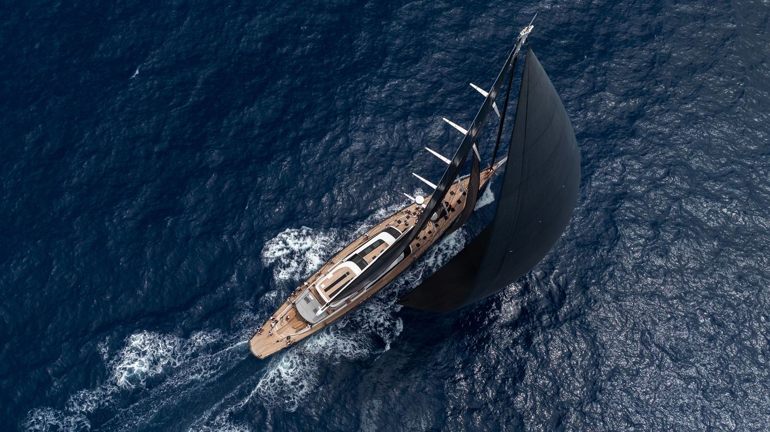 Aerial view of a large naval vessel sailing through dark blue ocean waters leaving a white wake trail behind.
