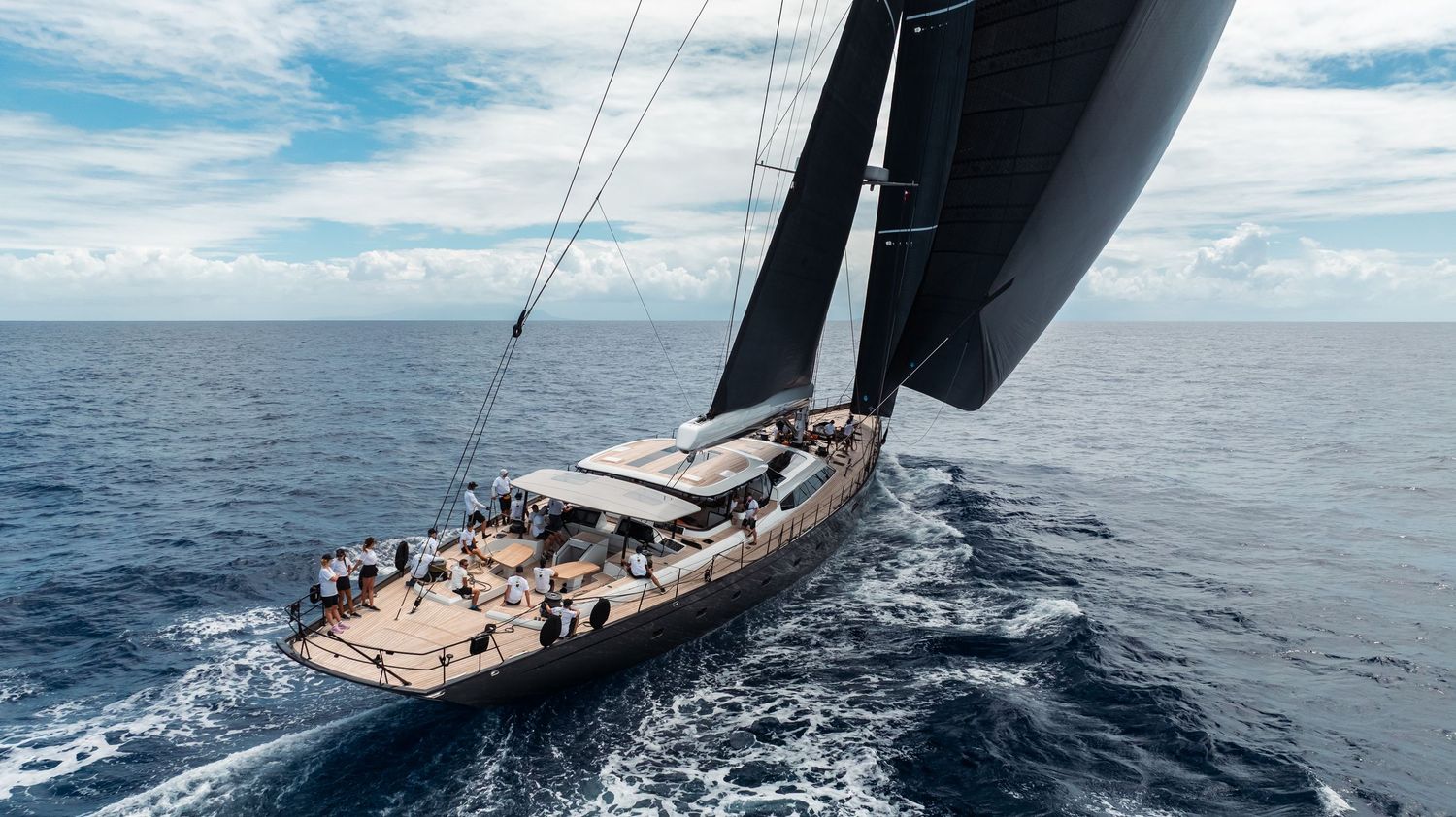 A luxury sailing yacht glides through blue ocean waters with black sails against a cloudy sky.