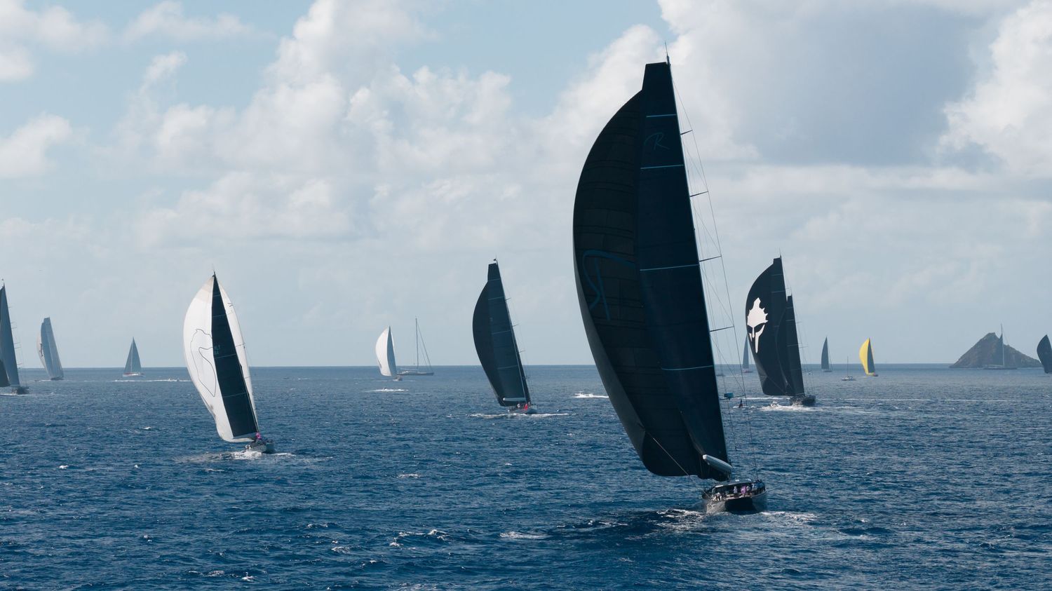 Fleet of sailboats with black spinnaker sails competing in a regatta on tropical waters.