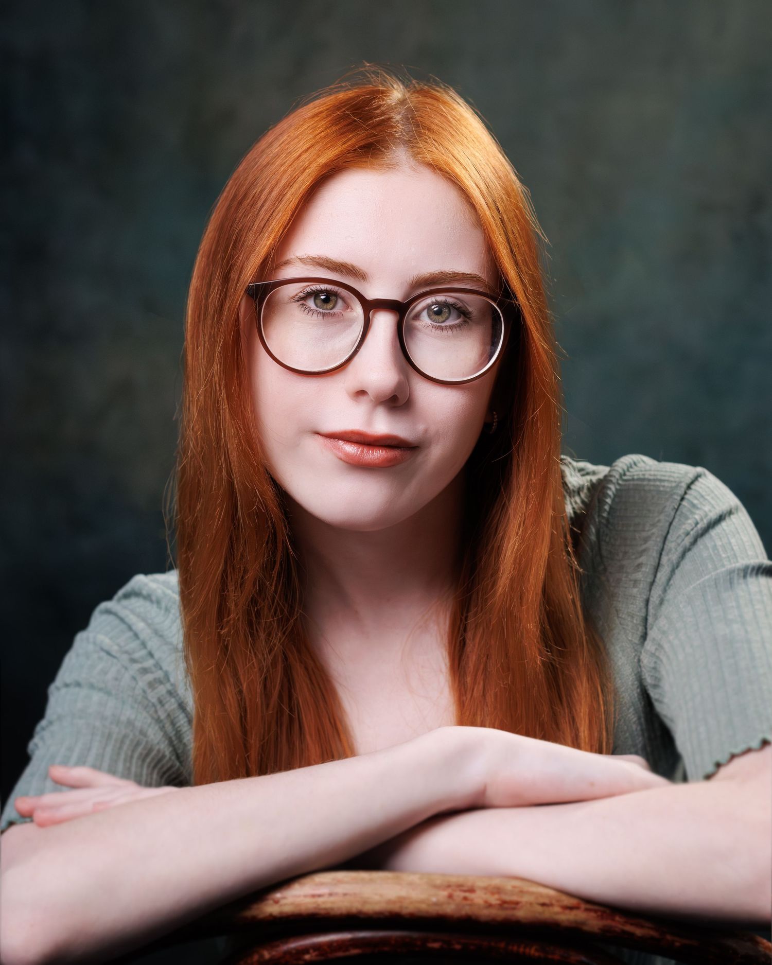 Portrait of someone with long red hair and glasses wearing a grey sweater against a dark background.