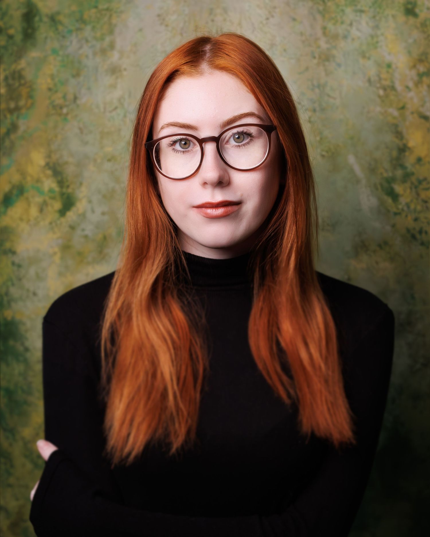 Portrait against textured green wall showing a person with red hair wearing black turtleneck and round glasses.
