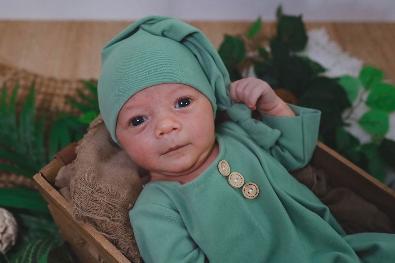 A baby wearing a green hat and outfit lies on a brown blanket surrounded by green leaves and foliage.