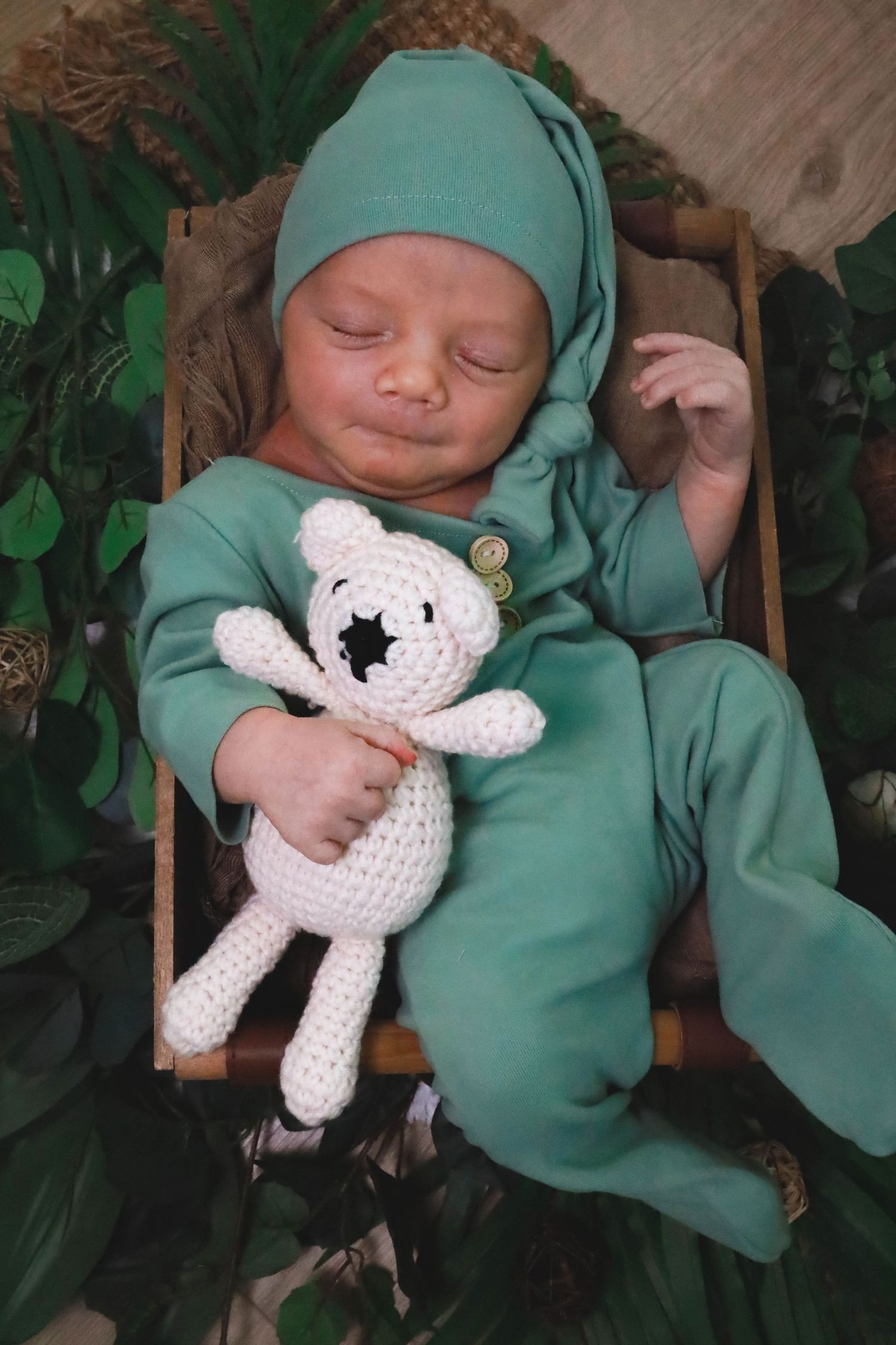 A sleeping baby in green clothing holds a small white crocheted stuffed animal toy.