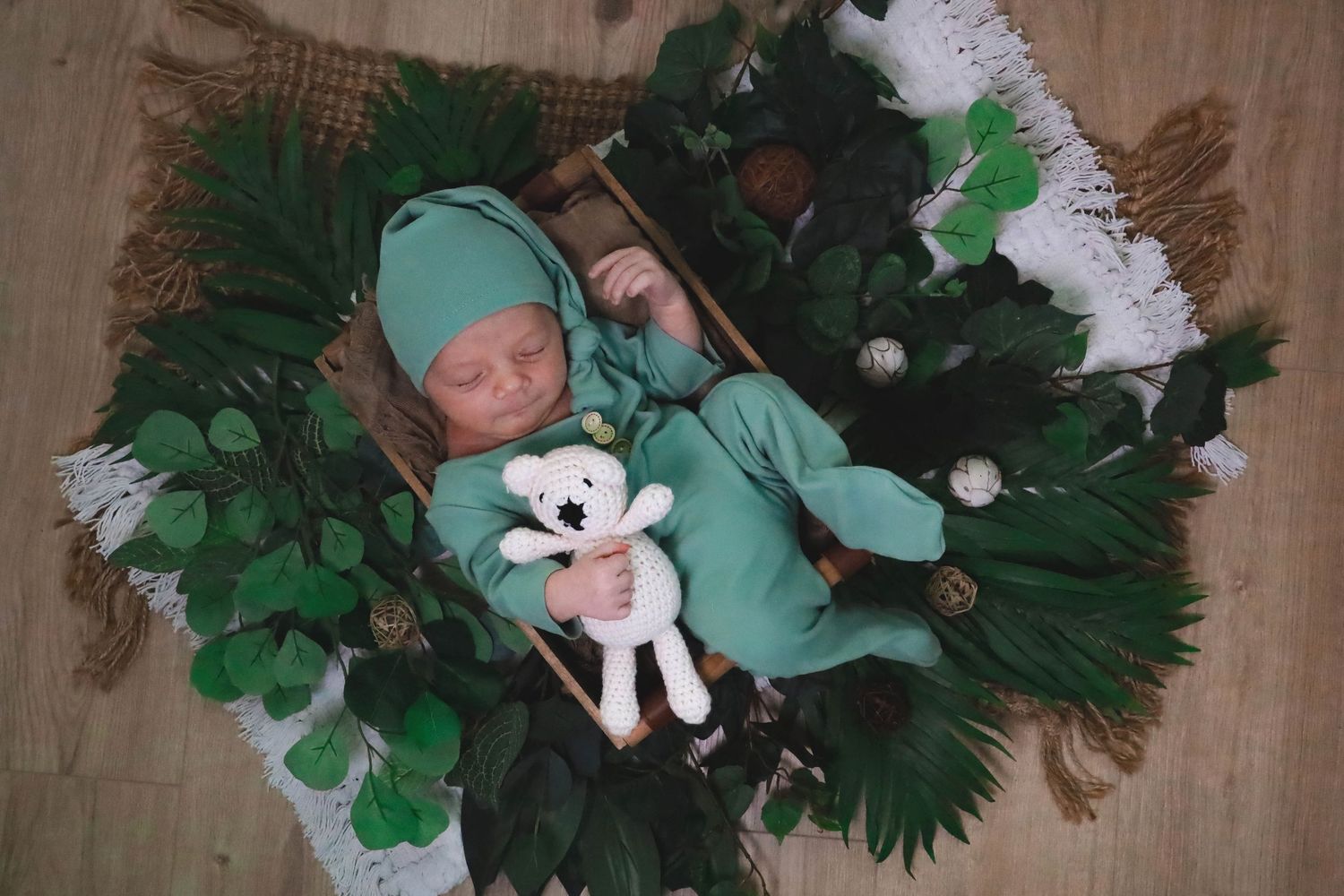 A small white plush toy nestled among green artificial monstera leaves and tropical foliage decorations.