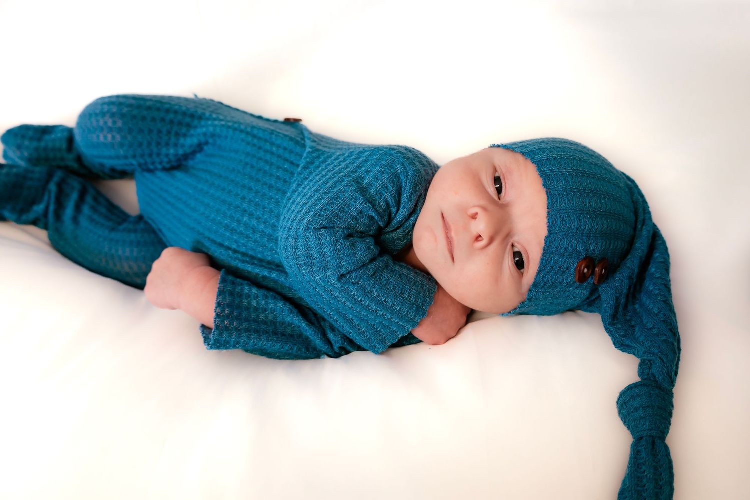 A baby in a teal knit romper lies on a white background showing different sleeping positions.