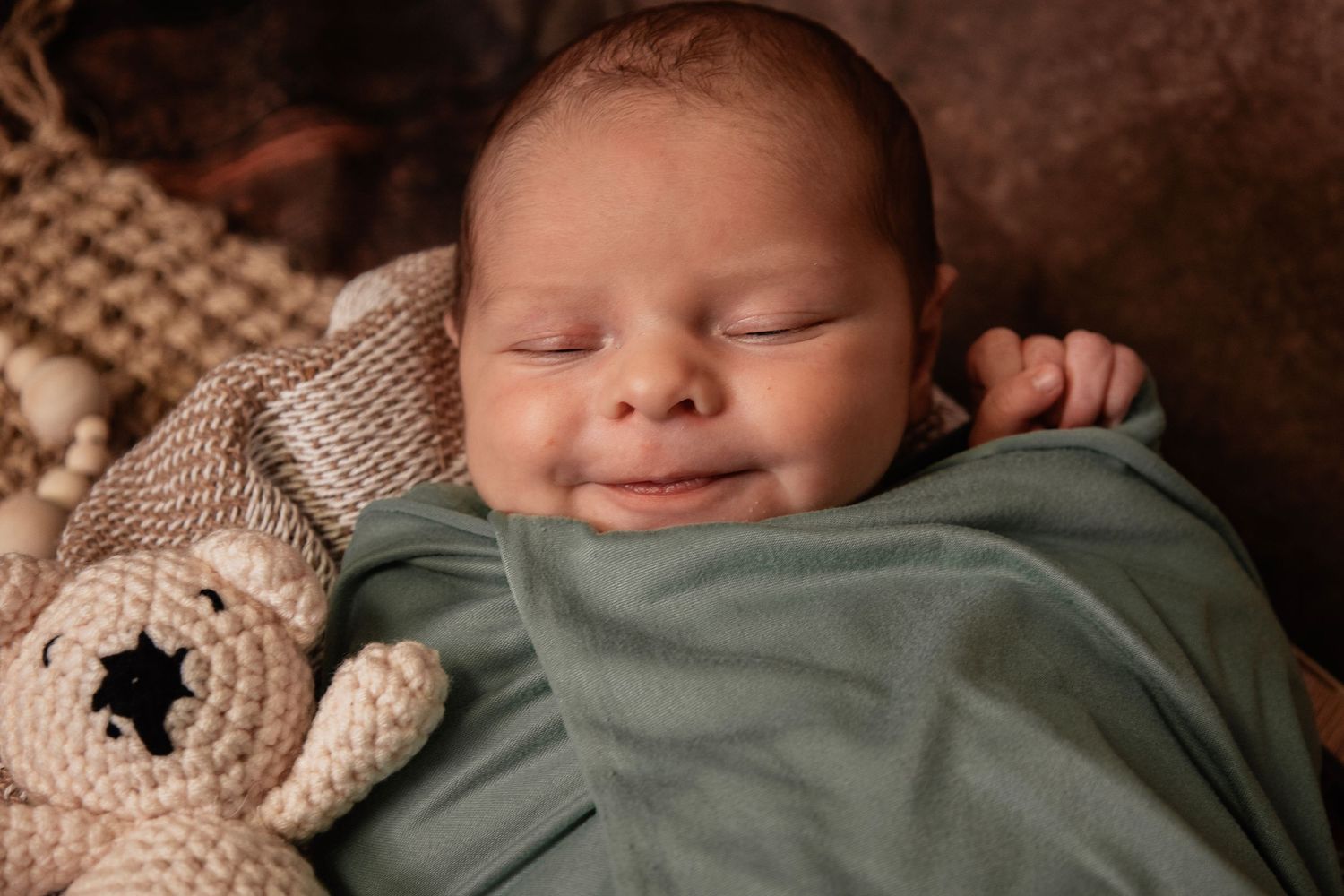 A sleeping newborn wrapped in a cozy knit blanket lies peacefully against a warm brown backdrop.