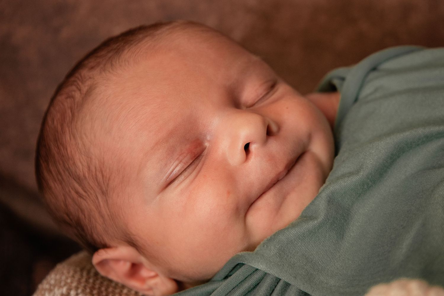 A sleeping baby in a green swaddle blanket lies peacefully on a soft surface.