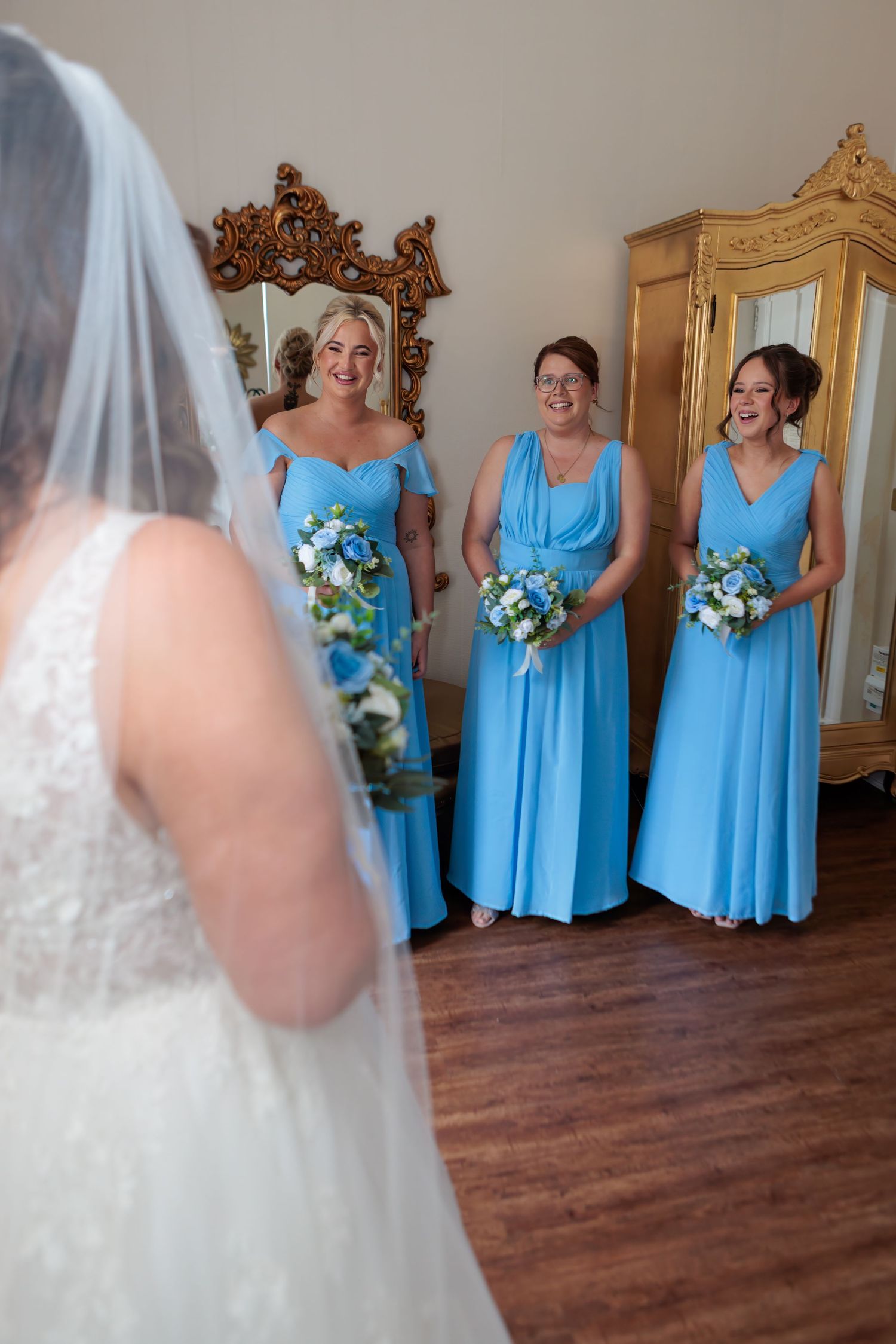 Wedding party members in light blue bridesmaid dresses holding blue and white bouquets stand together on hardwood floors.