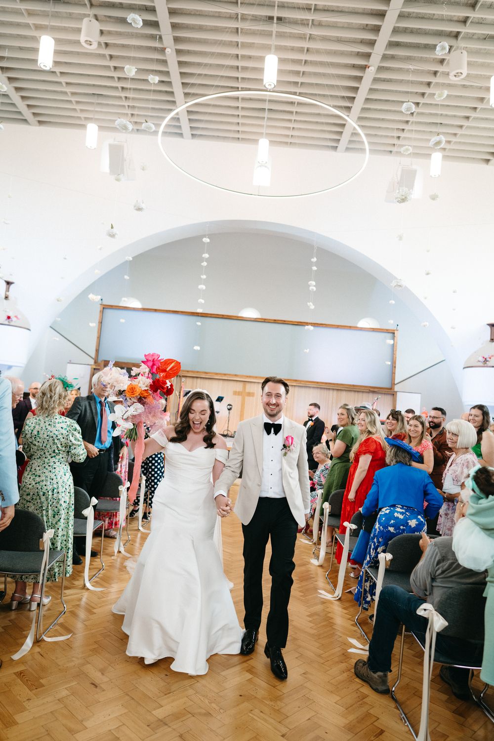 Wedding reception guests dance and celebrate in a bright white ballroom with clothing racks visible along the walls.