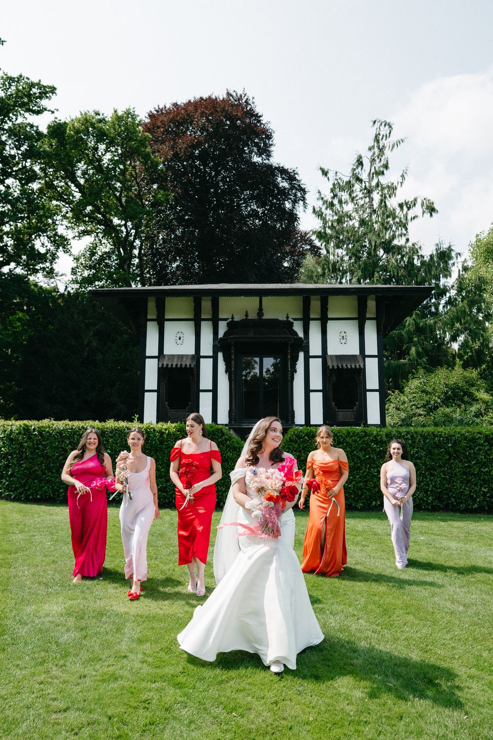 A wedding party poses on a manicured lawn in front of a black and white garden pavilion on a sunny summer day.