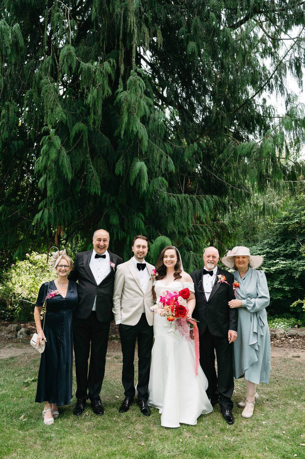 Wedding party poses together outdoors under evergreen trees with pink bouquet and formal attire.