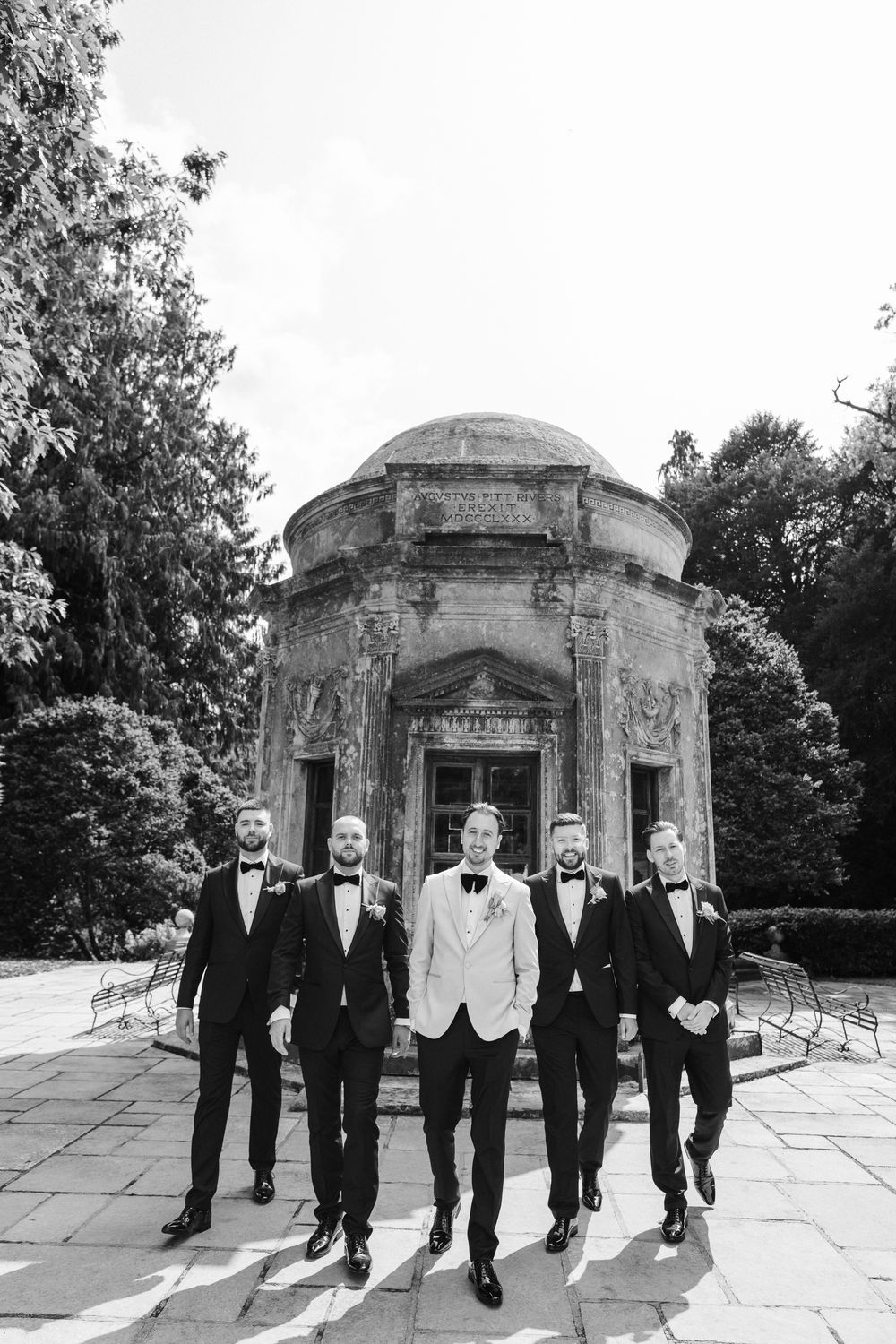 Groomsmen in formal black and white attire walk together in front of a stone gazebo structure. Taken by modern wedding photographer.