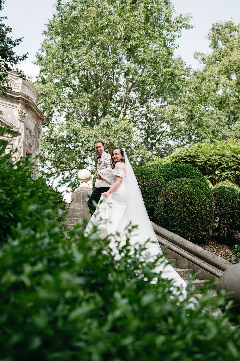 A couple in wedding attire poses elegantly on stone steps surrounded by lush green landscaping and boxwood shrubs.