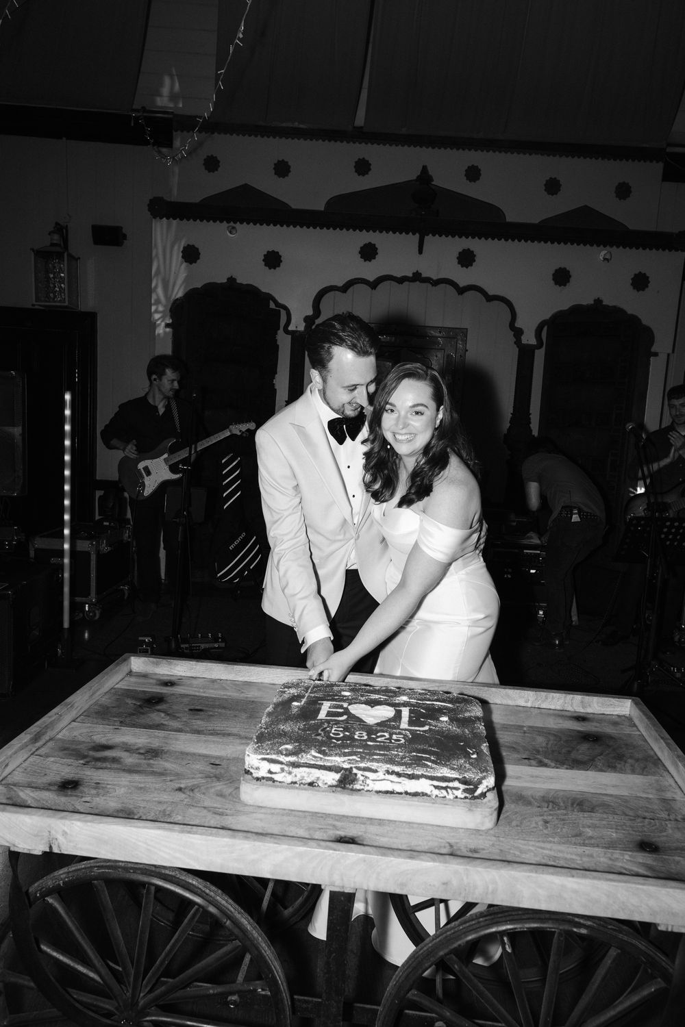 A couple in formal wedding attire cuts their cake together at a rustic wooden table in black and white photography.