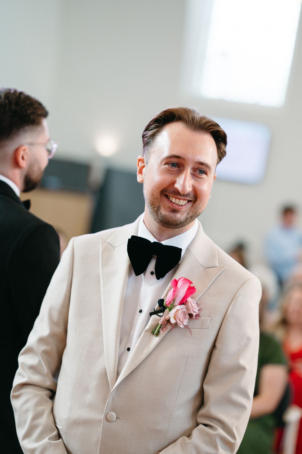 A groom in a cream-colored suit and black bowtie smiles during a wedding ceremony. Taken by modern editorial wedding photographer.