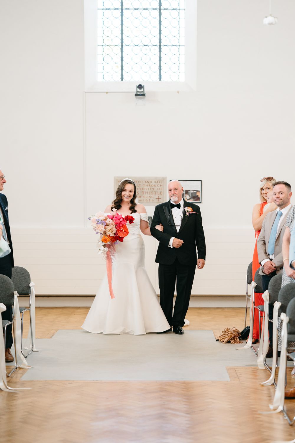 Elegant wedding ceremony in a bright white room with coral accents and guests seated in chairs along the aisle.