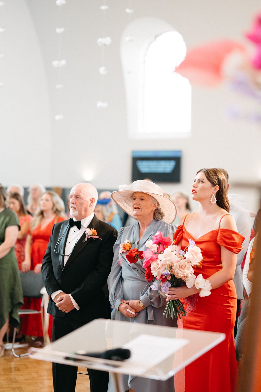 Wedding guests in formal attire including a tuxedo and red dress stand together during a ceremony in an elegant venue.