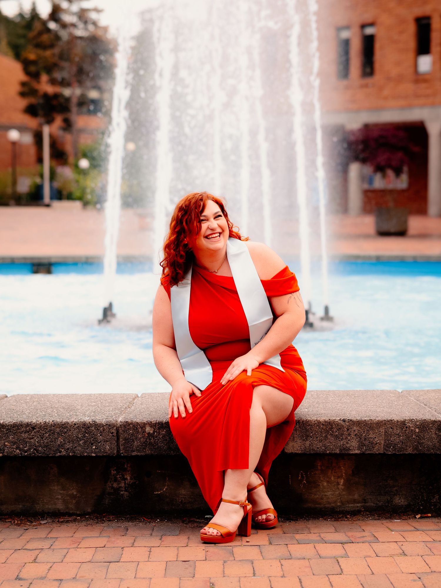 A person in a vibrant red dress sits by a fountain on a brick patio, with water spraying in the background.