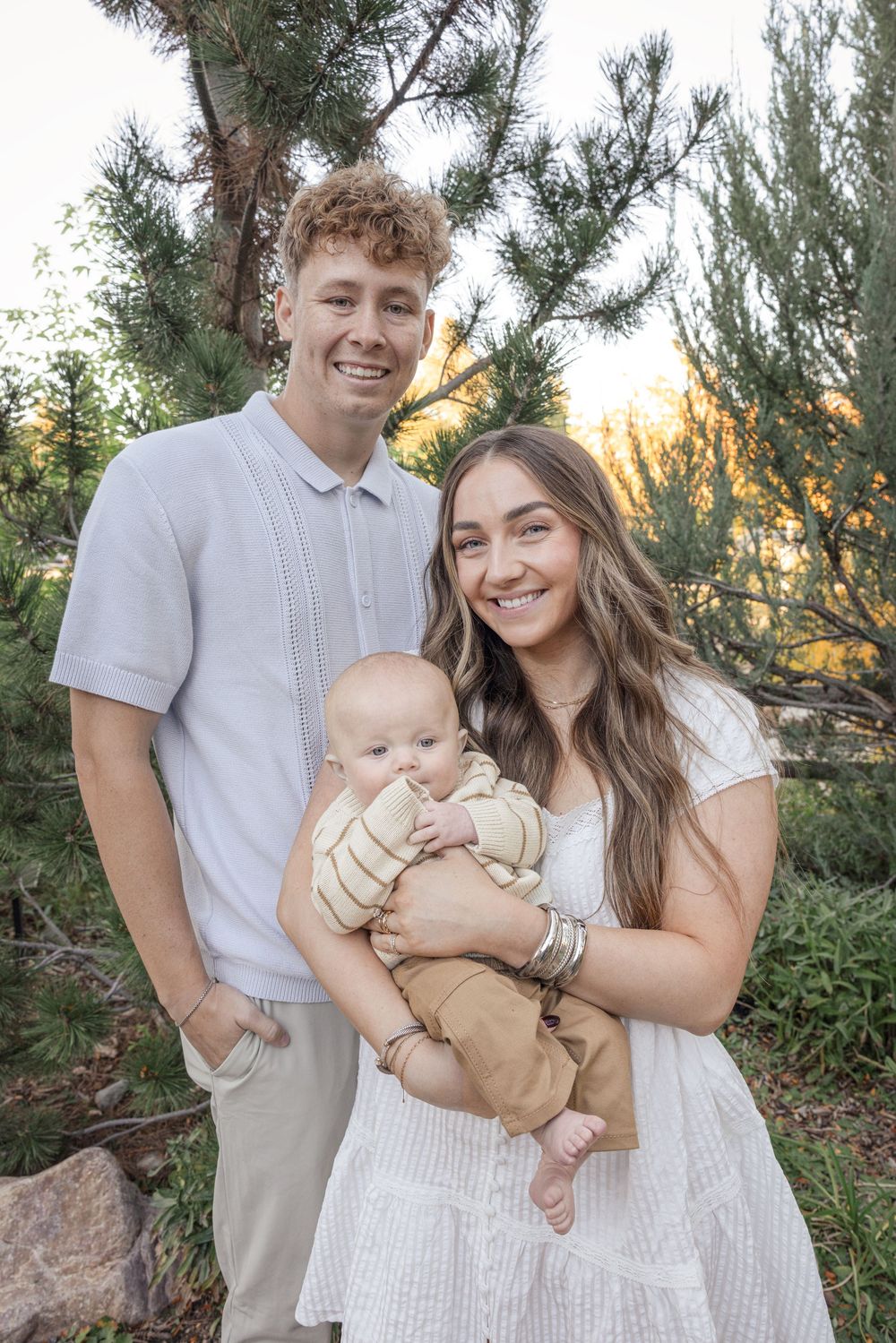 A family in coordinating white and beige outfits poses together outdoors at sunset among pine trees.