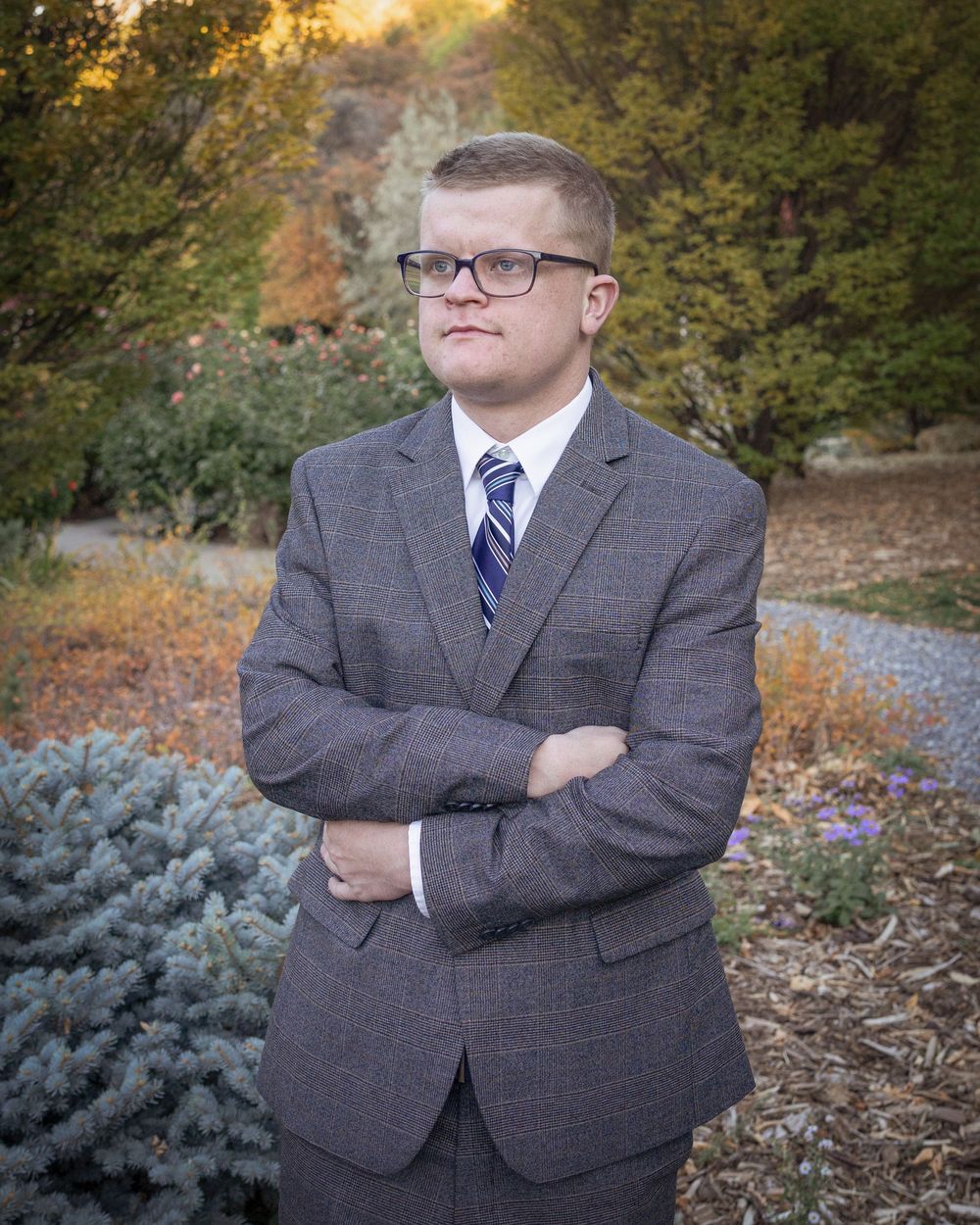 A person in a gray suit and blue tie poses with crossed arms in an autumn outdoor setting with trees and shrubs.