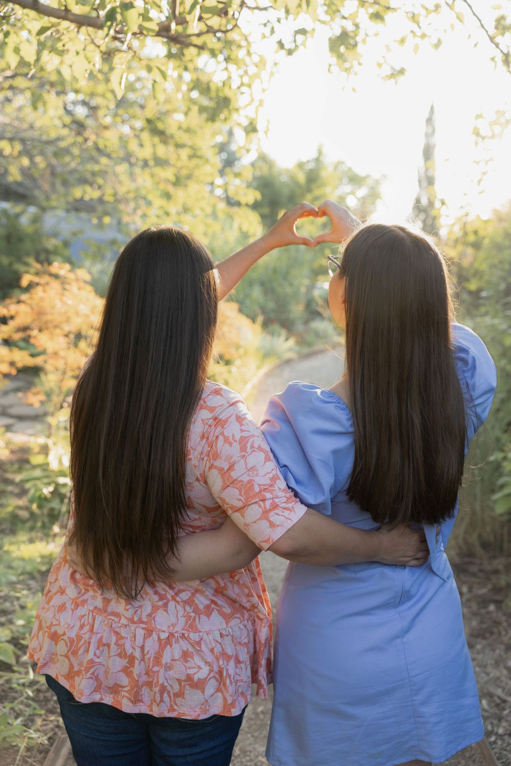 Two people with long dark hair making heart shape with hands against golden sunset.
