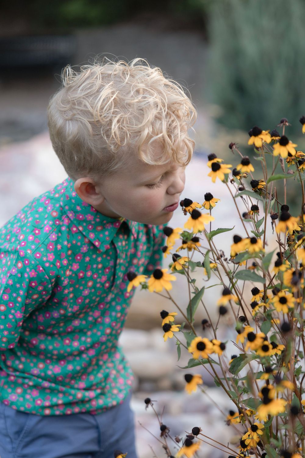 A small child in a polka dot shirt leans in to smell yellow wildflowers in a garden setting.