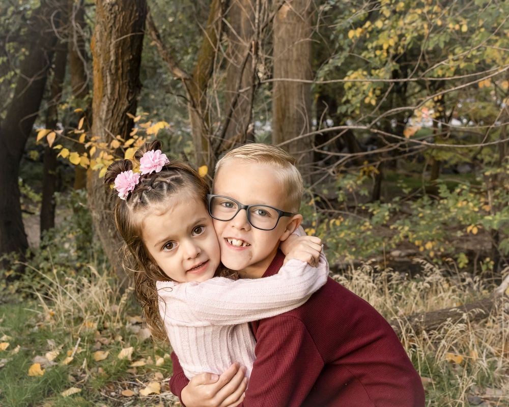 Two people in cozy sweaters embrace while surrounded by autumn trees and fallen leaves in a natural outdoor setting.