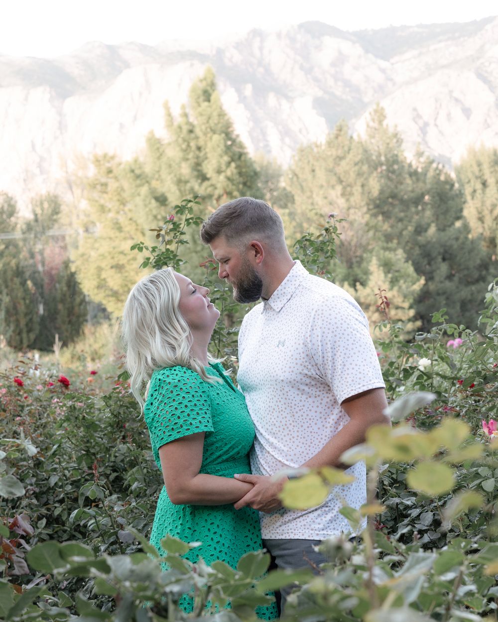 A couple embraces in a garden with red roses and snow-capped mountains in the background.